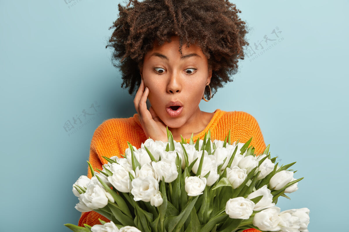 理发惊讶的美国黑人女士盯着白色美丽的花朵 不敢相信的眼睛 手放在脸颊上 穿着橙色毛衣 隔着蓝色的墙人们和意想不到的反应概念花女人情感