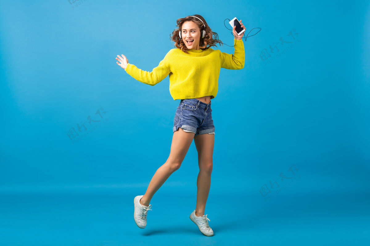 长度迷人的笑容可掬的快乐女人戴着耳机跳着舞听着音乐 穿着时髦的服装 隔离在蓝色的工作室背景下 穿着短裤和黄色的毛衣音乐牛仔裤听