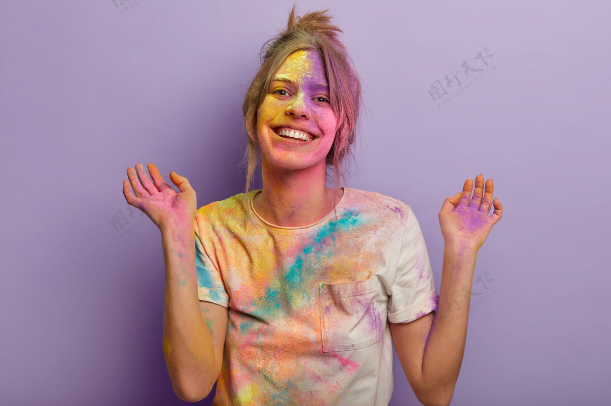 传统快乐的欧洲女人 表情无忧无虑 举手 涂上五颜六色的染料 穿着白色t恤 高兴地微笑 庆祝节日 把派对隔离在紫色的墙上积极欣喜若狂牙齿