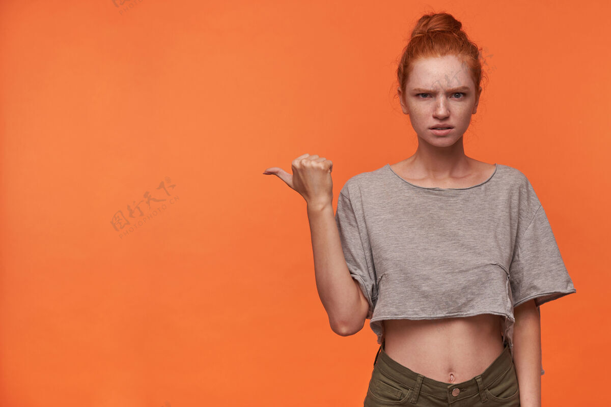 拇指一幅严肃的年轻女读者的画像 她梳着发髻 皱着眉头对着镜头 举起手 拇指指向一边 站在橙色背景上T恤头发短裤