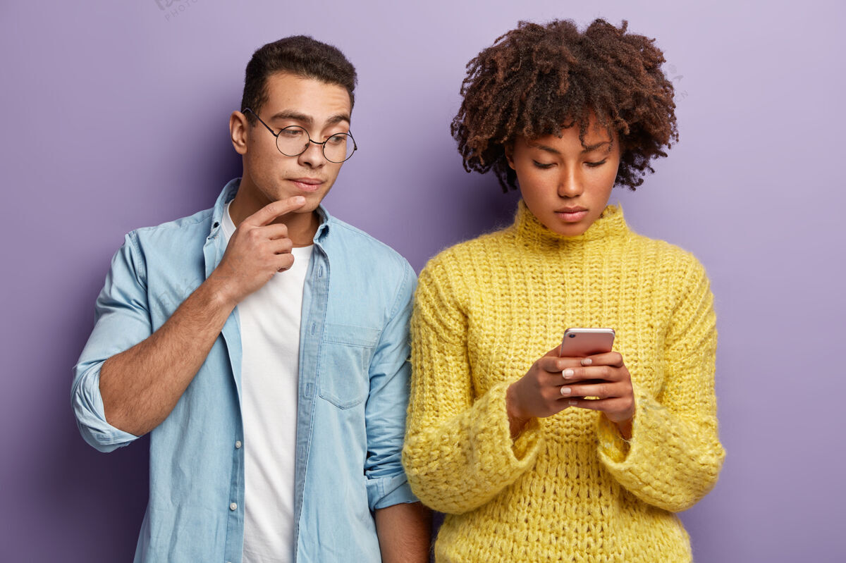 非洲不同的女人和男人站在室内 深色皮肤的女士专注于智能手机 输入反馈 发送短信 好奇的家伙偷看屏幕 穿着牛仔衬衫多种族关系概念小玩意年轻女朋友