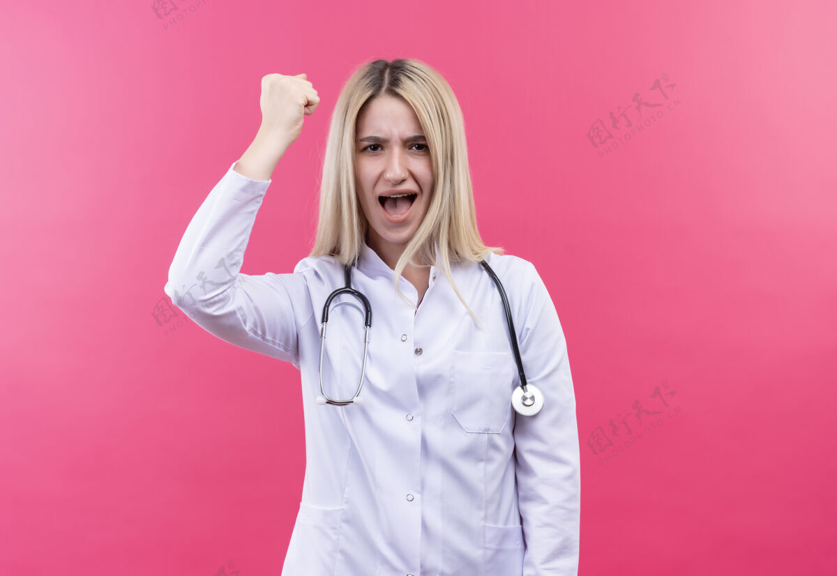 粉红色严格的医生年轻的金发女孩穿着医用长袍手持听诊器在孤立的粉红色背景上金发穿着女孩