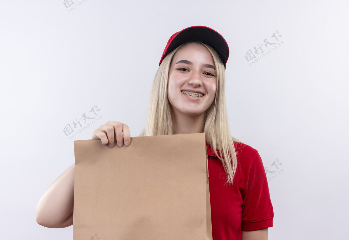支架带着微笑的小女孩穿着红色t恤 戴着帽子 戴着牙套 在孤立的白色背景上拿着纸袋白色年轻送货