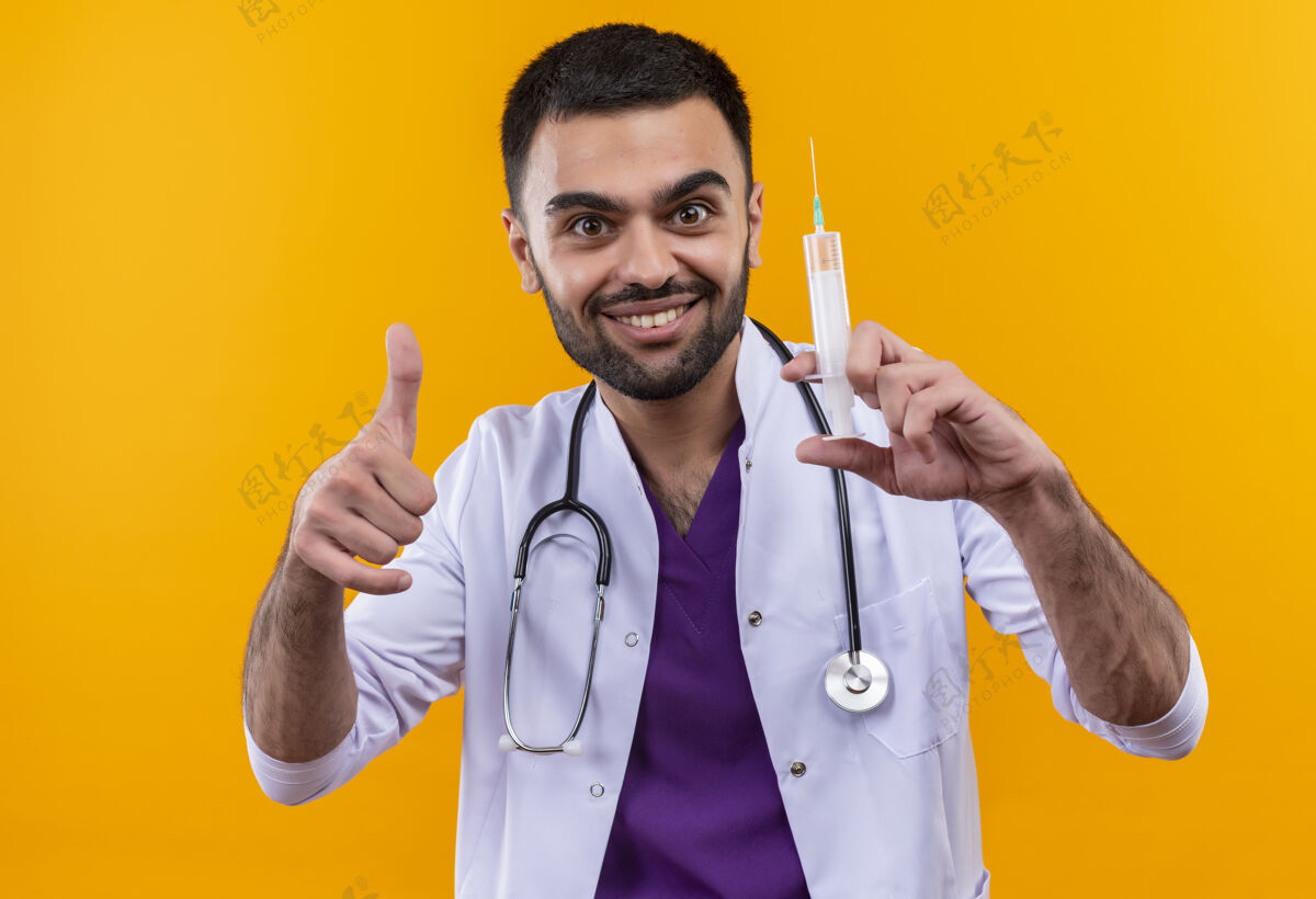 向上微笑着的年轻男医生穿着听诊器医用长袍 拿着注射器 他的大拇指向上竖立在孤立的黄色背景上年轻微笑医疗