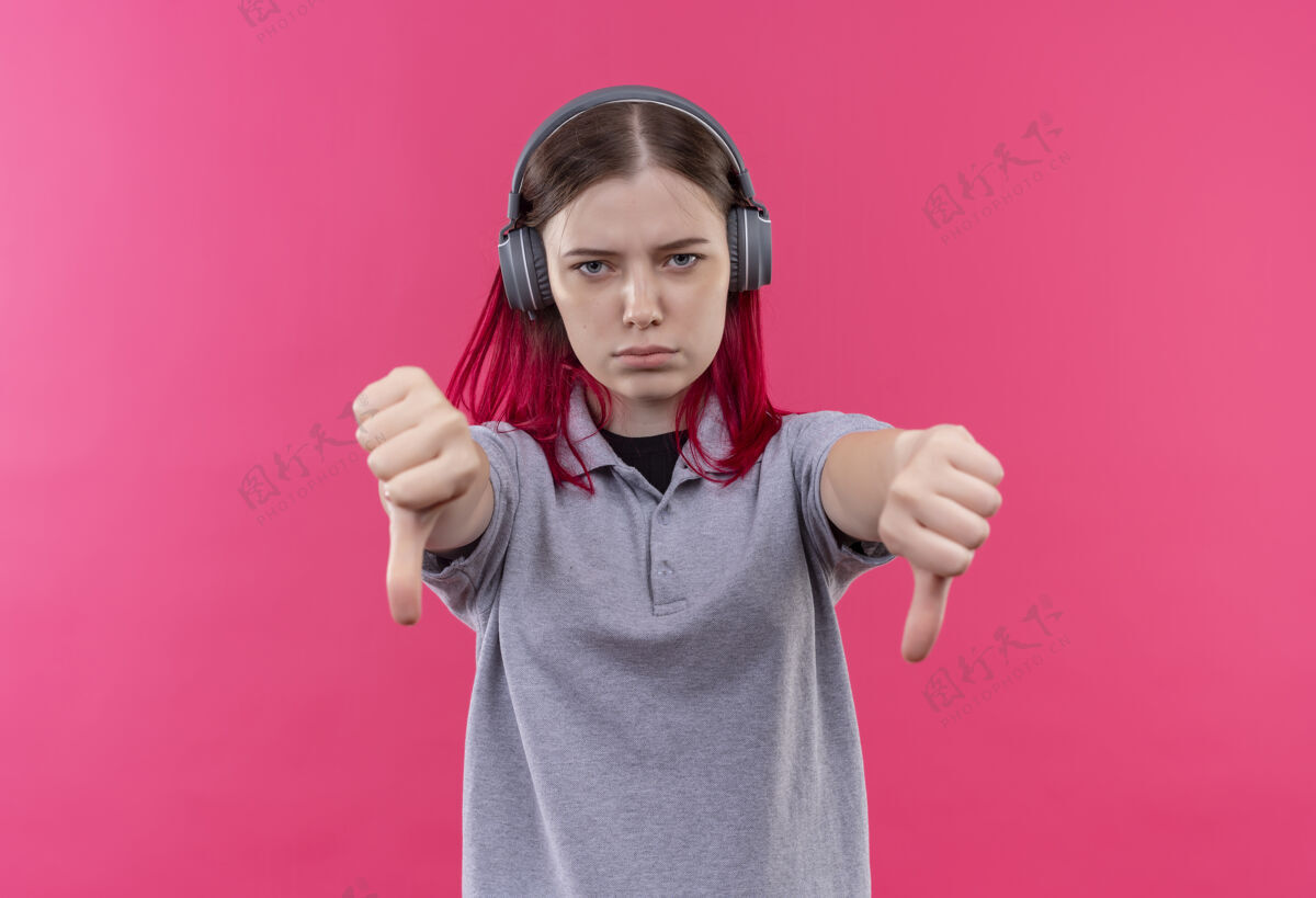 灰色戴着耳机 穿着灰色t恤的年轻漂亮女孩在孤立的粉色背景上竖起大拇指年轻粉色女孩