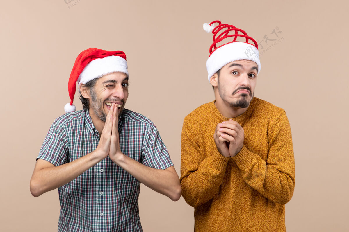 搞笑正面图两个戴着圣诞帽的滑稽家伙一个在米色孤立背景上眨着眼睛前面眨眼男性