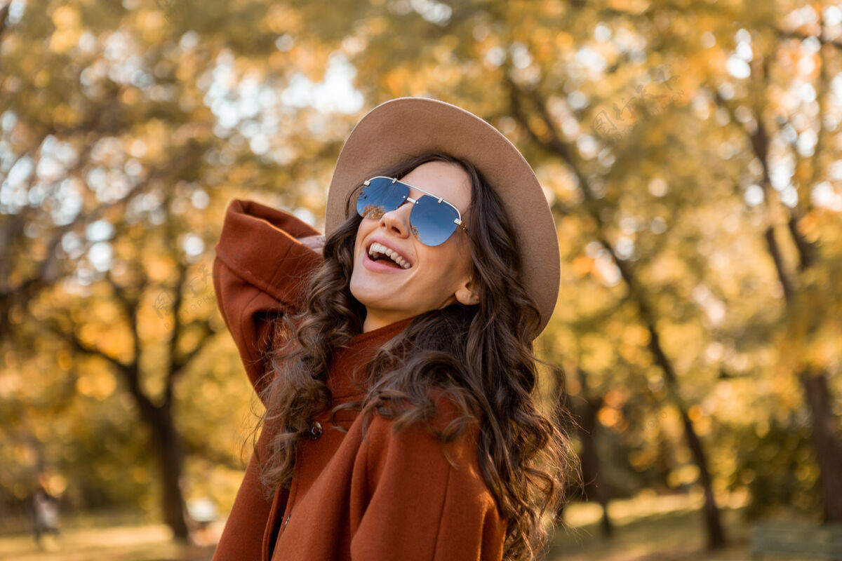 漂亮迷人的时尚微笑的女人卷发漫步在街头公园穿着温暖的棕色外套秋季流行时尚 街头风格戴帽子和太阳镜优雅服装休闲