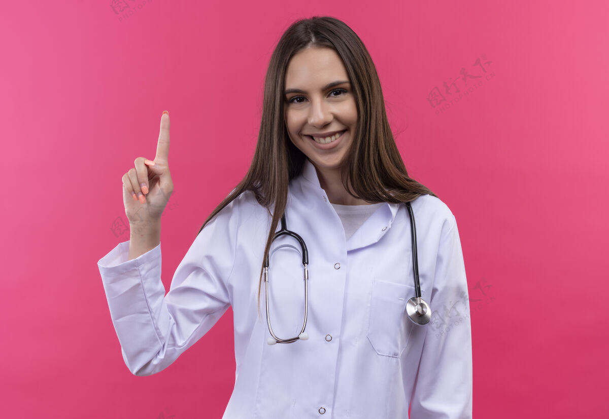 长袍微笑的年轻医生女孩穿着听诊器医用长袍 手指指向孤立的粉红色背景上听诊器微笑医疗