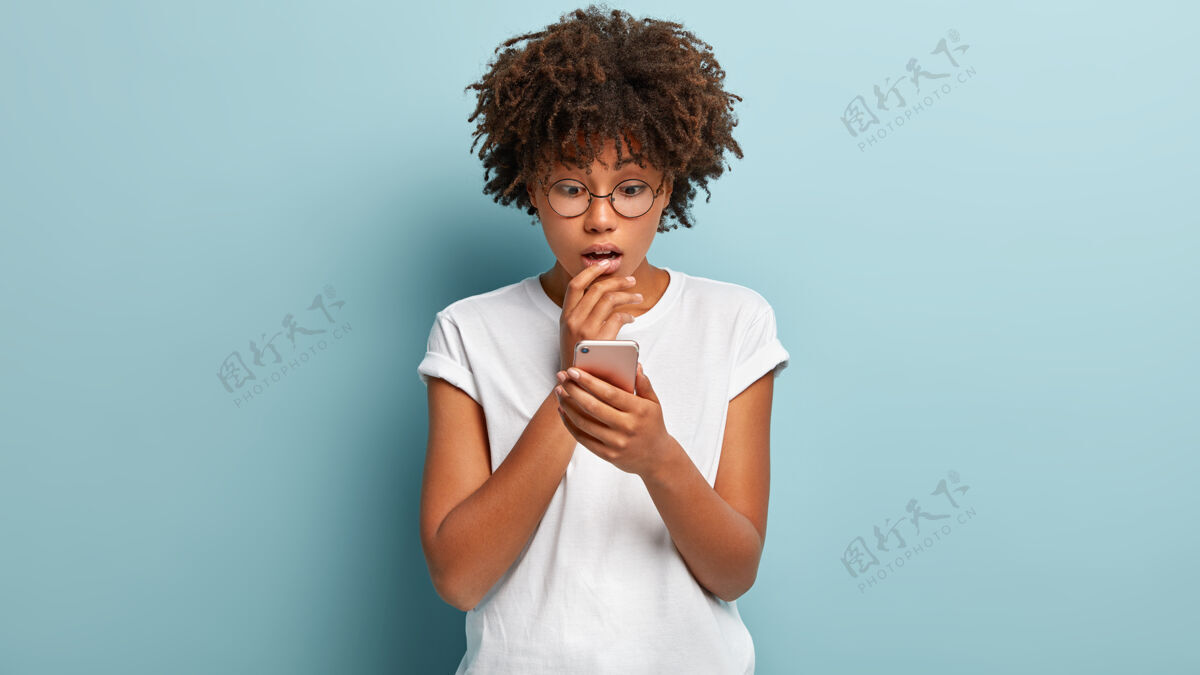 浏览在室内拍摄的震惊的非洲裔美国妇女集中在手机 阅读惊人的可怕消息 使用现代应用程序 戴着视力矫正眼镜隔离在蓝色墙壁错过了电话应用程序短信在线
