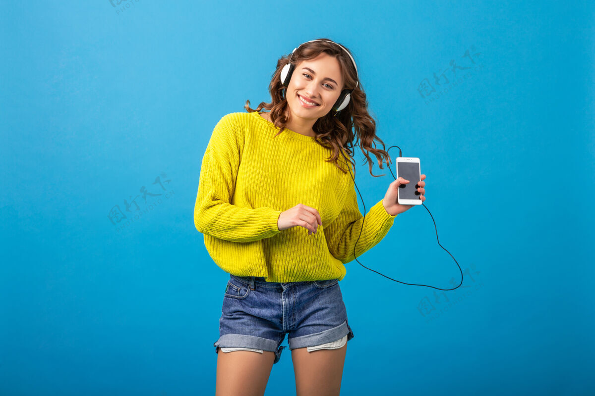运动迷人的笑容可掬的快乐女人戴着耳机跳舞听音乐 穿着时髦的服装 隔离在蓝色的工作室背景下 穿着短裤和黄色毛衣跳跃音乐耳机
