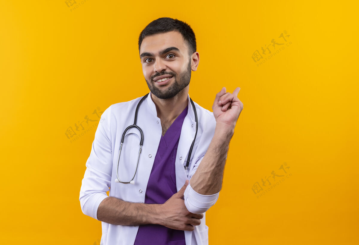听诊器高兴的年轻男医生穿着听诊器医用长袍 用手指指着背上孤立的黄色背景医学穴位背部