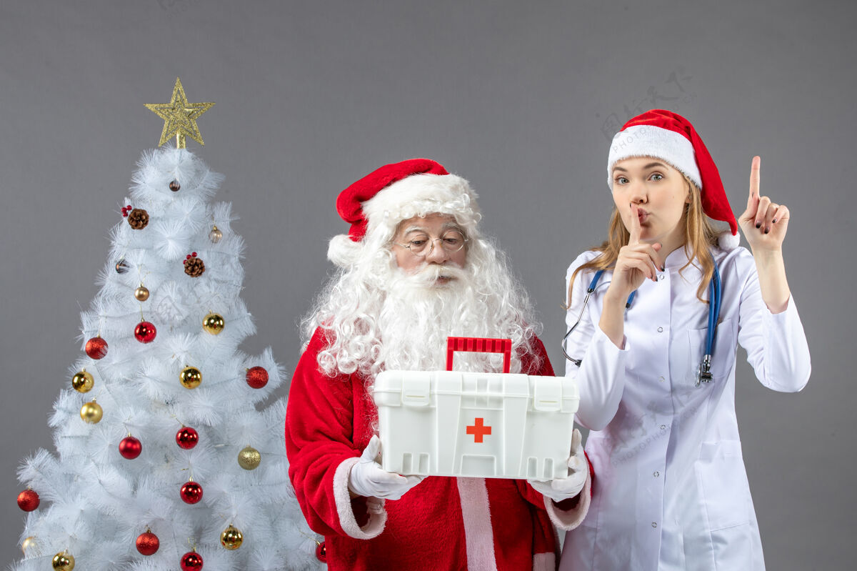 帽子圣诞老人和给他急救箱的女医生在灰色墙上的正视图工具包十二月谁