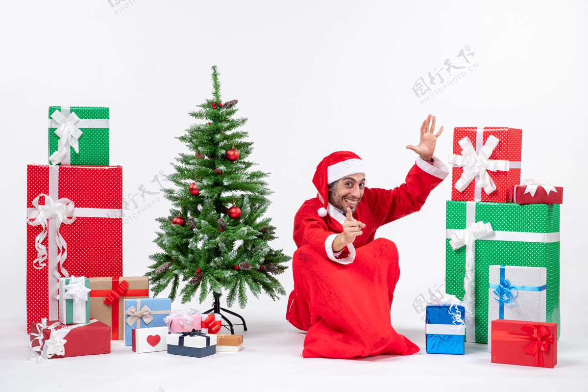 圣诞老人笑容满面的年轻人打扮成圣诞老人 拿着礼物和装饰好的圣诞树坐在地上 在白色背景上指着前方的东西装饰向前礼物