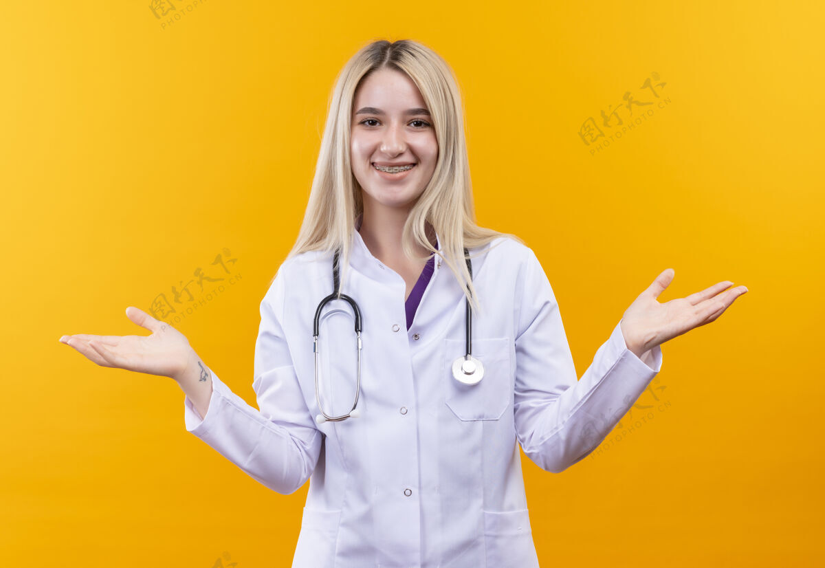 黄色微笑的医生穿着医用长袍戴着听诊器和牙套的年轻女孩在孤立的黄色背景上展开双手年轻牙科医生