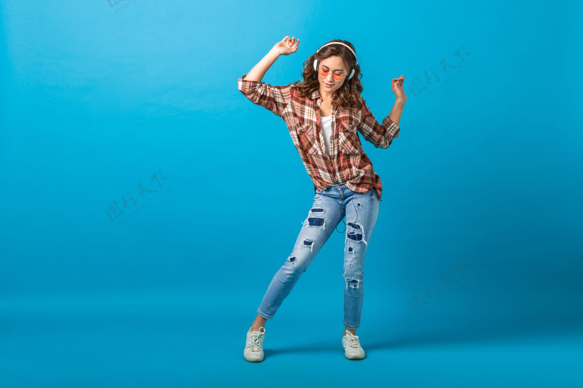 耳机迷人的快乐女人摆出欢快的姿势 戴着耳机 穿着格子衬衫和牛仔裤 在蓝色的工作室背景下 听着音乐 抬头仰望饱满女性听