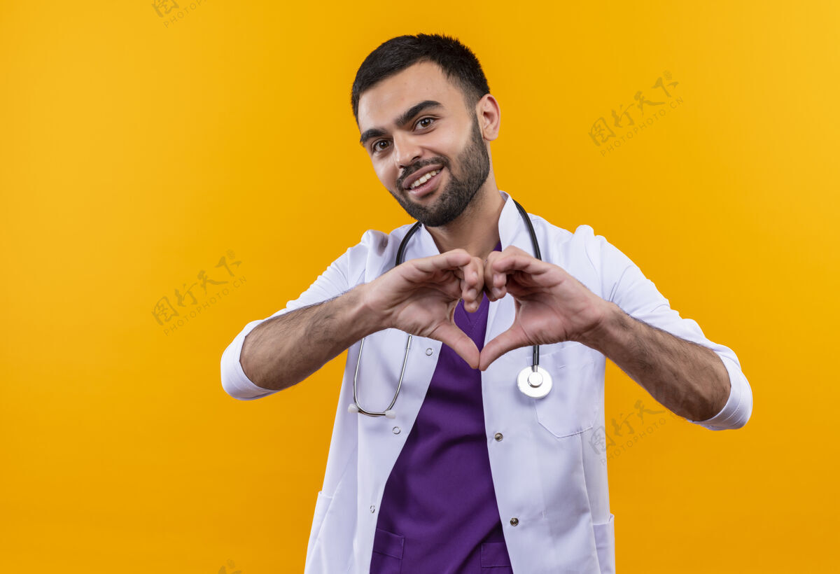 黄色高兴的年轻男医生穿着听诊器医用长袍 在孤立的黄色背景上显示心脏姿态医学男性礼服