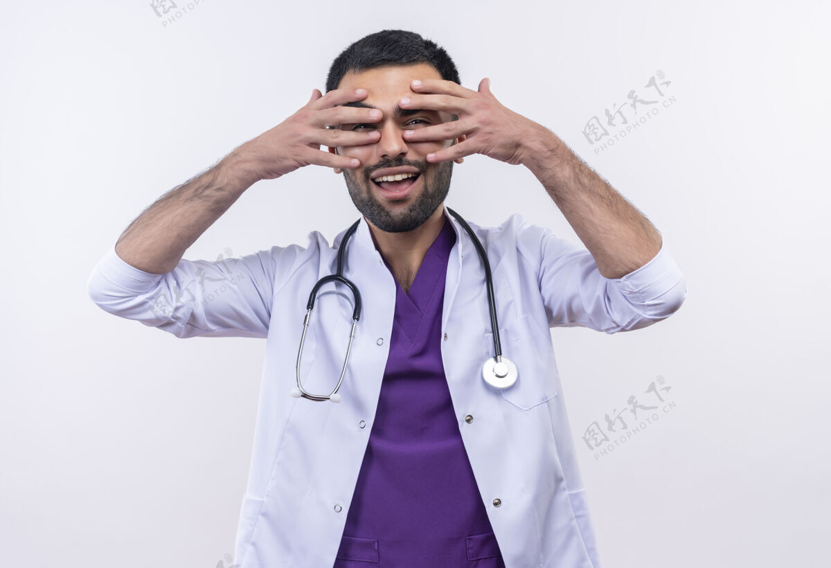 男性微笑的年轻男医生穿着听诊器医用长袍 双手放在孤立的白色背景上的眼睛上医疗年轻医生