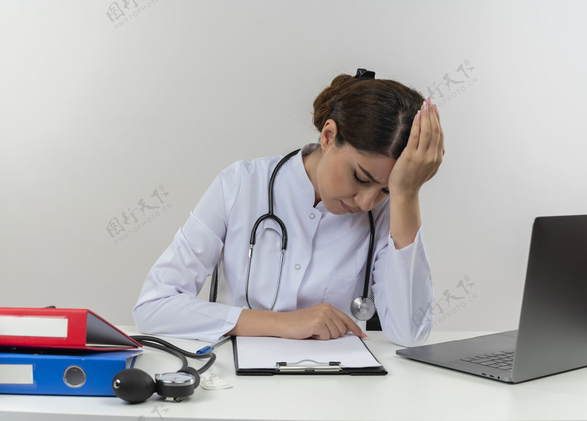 坐着疲惫不堪的年轻女医生穿着医用长袍 手持听诊器坐在办公桌旁 拿着医疗工具在电脑上工作 低头留着复印空间穿放下办公桌