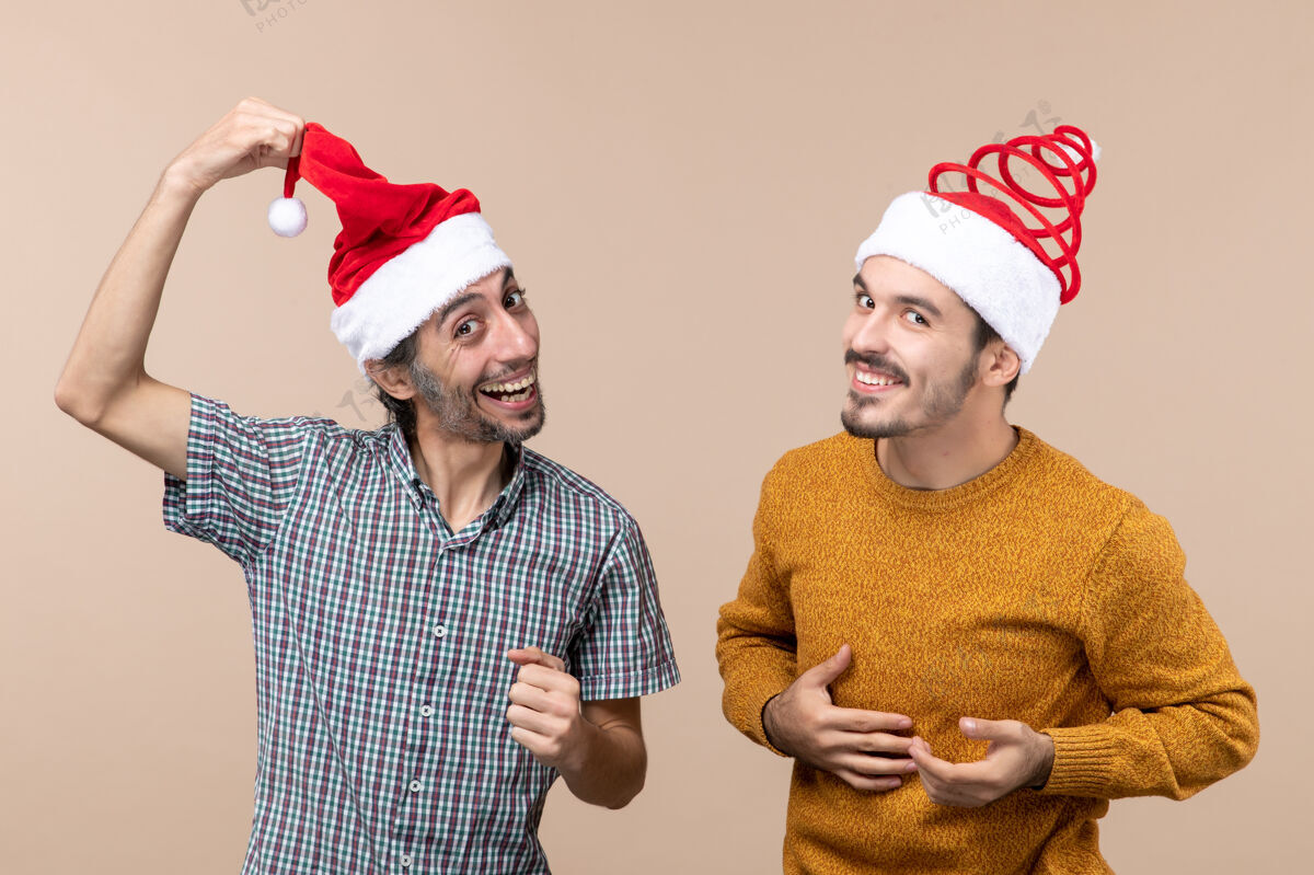 大笑正面图：两个戴着圣诞老人帽子的家伙 其中一个在米色孤立背景上展示他的帽子两个帽子两个大笑的家伙
