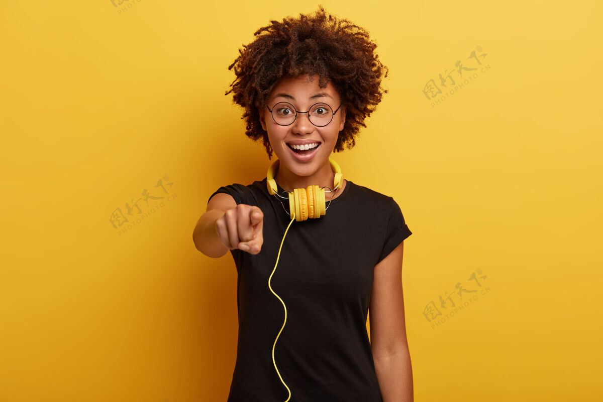 耳机可爱的积极的黑皮肤女孩穿着黑色t恤 圆眼镜 有黄色的耳机连接到一些小玩意 是真正的梅洛曼播放列表音频青少年
