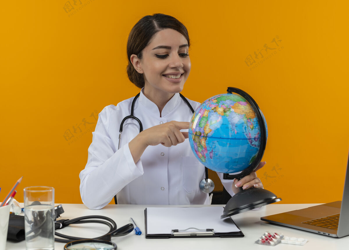 地球仪面带微笑的年轻女医生穿着医用长袍 手持听诊器坐在办公桌旁 手持医疗工具 将手指放在隔离黄色背景的地球仪上手指听诊器女