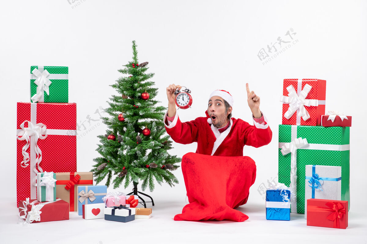 时钟惊喜的圣诞老人情绪激动地坐在地上 并显示附近的礼物和装饰圣诞树白色背景时钟圣诞树惊讶季节性