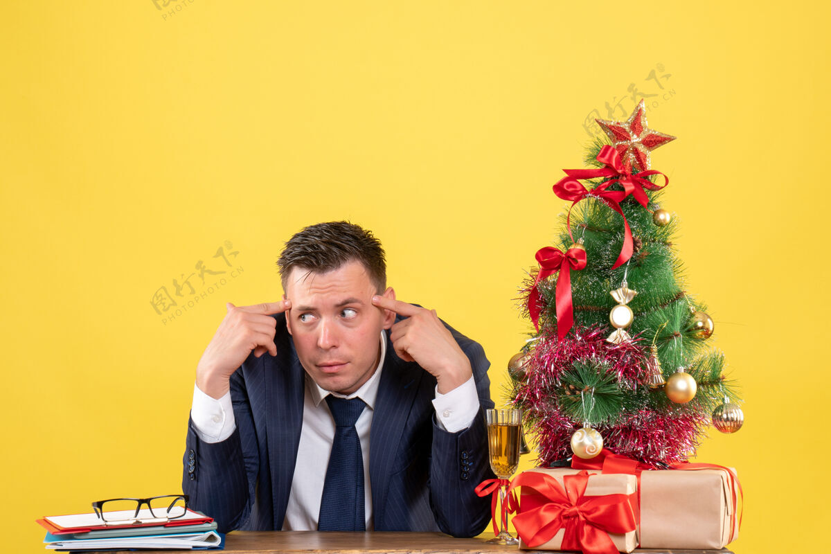 年轻人年轻人把手指放在他的太阳穴上 坐在圣诞树旁的桌子上 把礼物放在黄色的纸上套装黄色庙宇