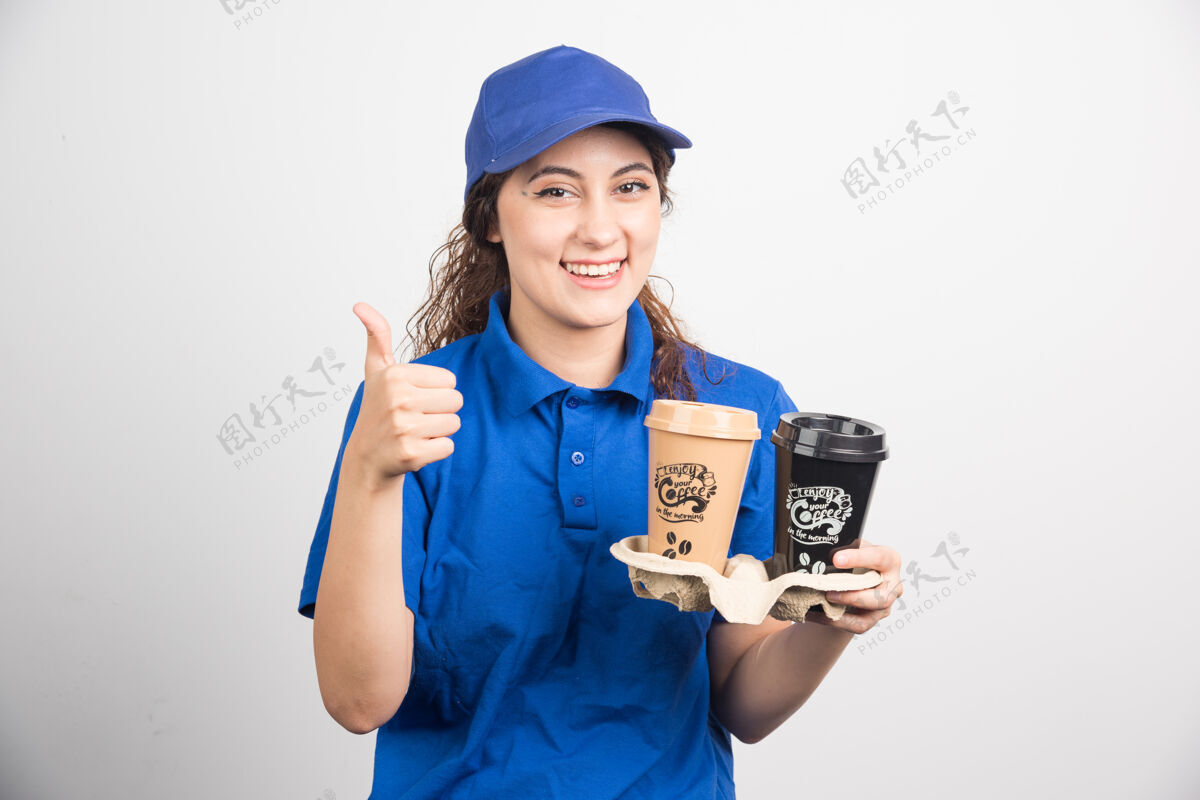 制服身着蓝色制服的女士举起大拇指 拿着两杯咖啡 背景是白色的高质量的照片帽子手女
