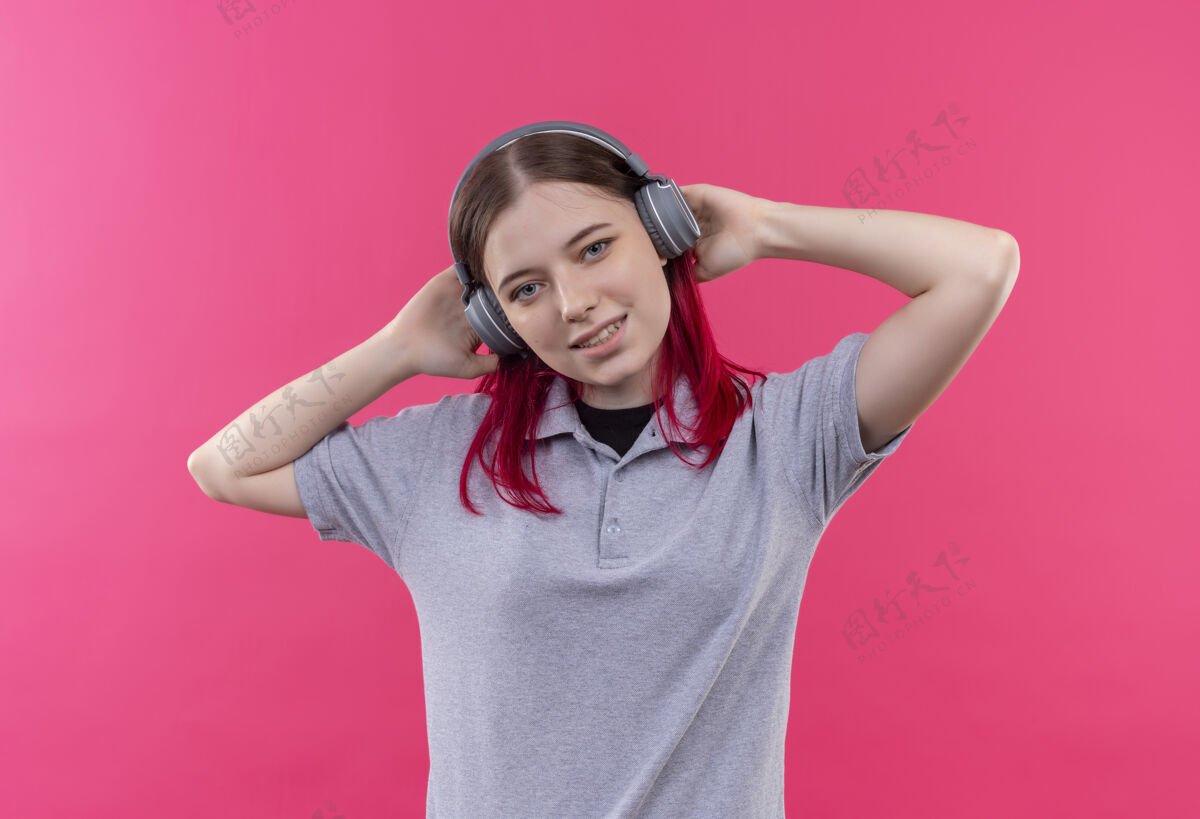 听高兴的年轻漂亮的女孩穿着灰色t恤听音乐耳机上孤立的粉红色背景穿耳机T恤
