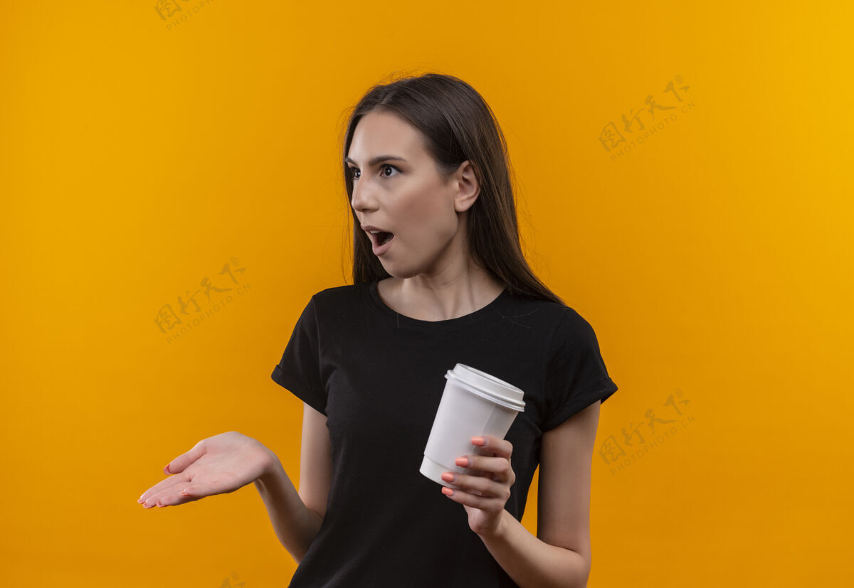 边上惊讶的年轻白人女孩穿着黑色t恤 手里拿着一杯咖啡 一边看着孤立的橙色背景穿着黑色女孩