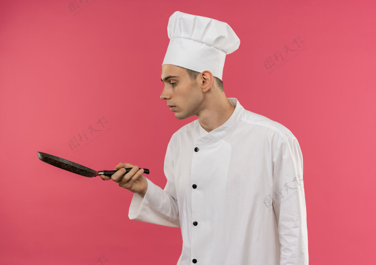 平底锅站在侧视图年轻的男厨师穿着厨师制服看着他手中的煎锅与副本空间穿厨师厨师