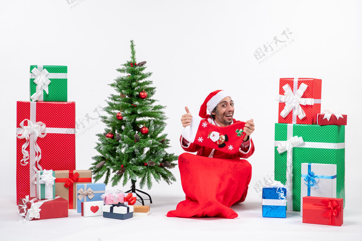 装饰喜庆的节日气氛 有趣的积极惊喜的圣诞老人坐在地上 展示圣诞袜子附近的礼物和装饰圣诞树上的白色背景圣诞老人圣诞老人积极的
