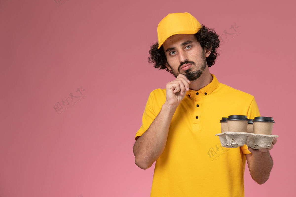 杯子正面图穿着黄色制服的男信使拿着棕色的咖啡杯 在粉红色的墙上思考思考工作视野