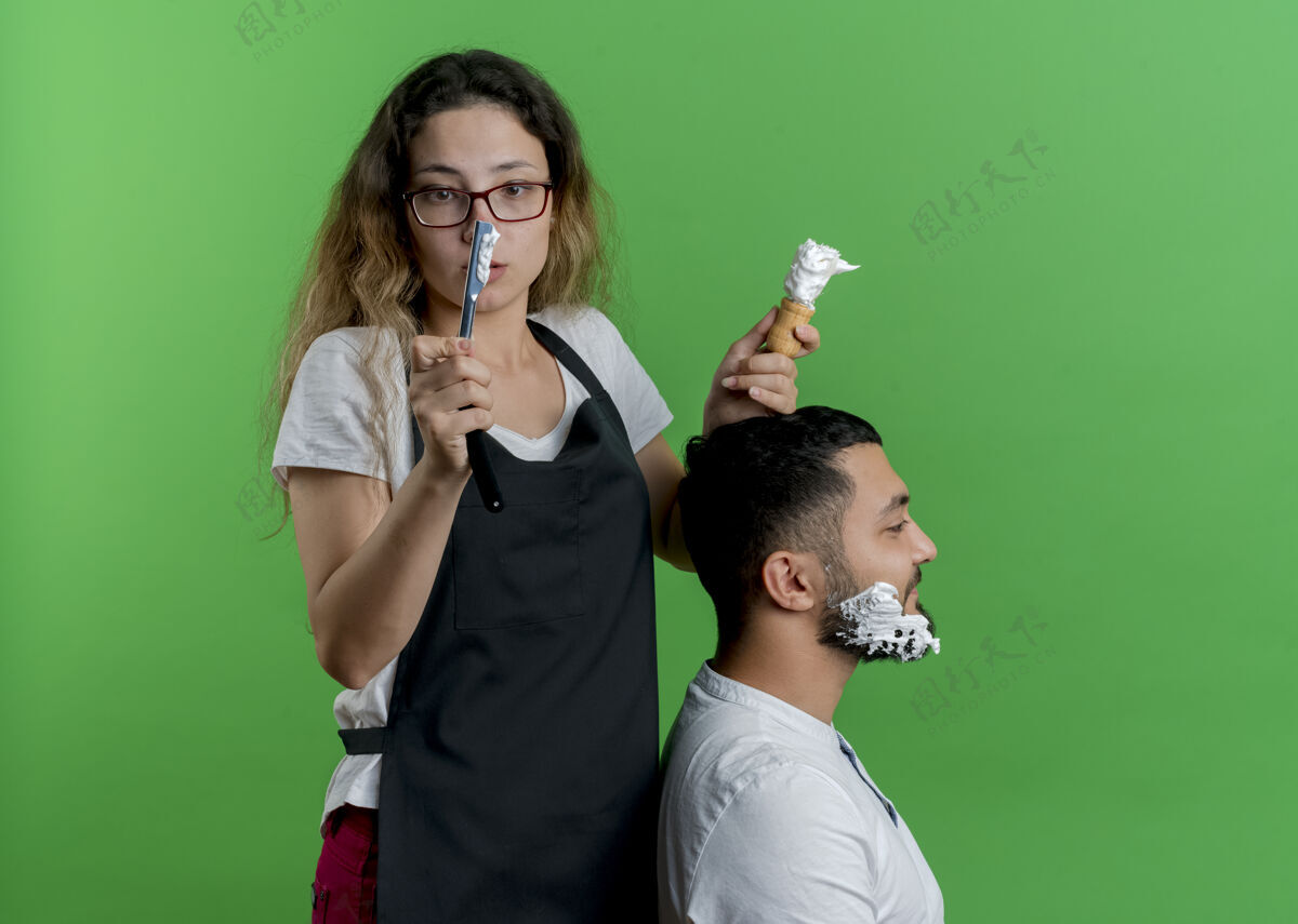 年轻年轻的专业美发师围裙里的女人把剃须泡沫和剃须的男人客户的胡子 很惊讶泡沫理发师剃须