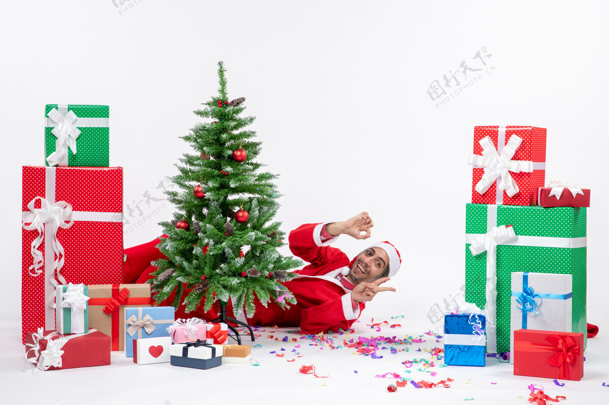 节日年轻的圣诞老人躺在圣诞树后面 在白色的背景下展示两件近乎礼物 这是节日的气氛冬青礼物圣诞老人