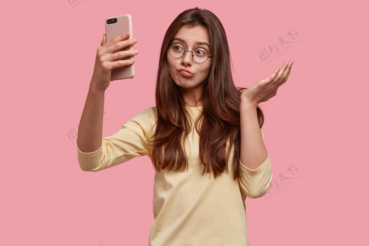 自拍漂亮犹豫不决的白人年轻女子形象以智能手机冷漠相看 自拍或打视频电话眼镜电子困惑
