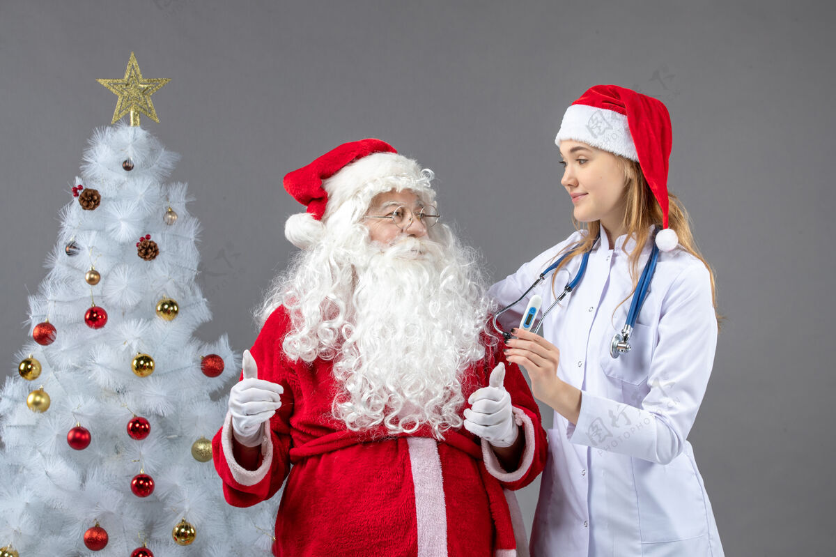 十二月圣诞老人和女医生的前视图 女医生拿着测温仪在灰色的墙上圣诞快乐护士人