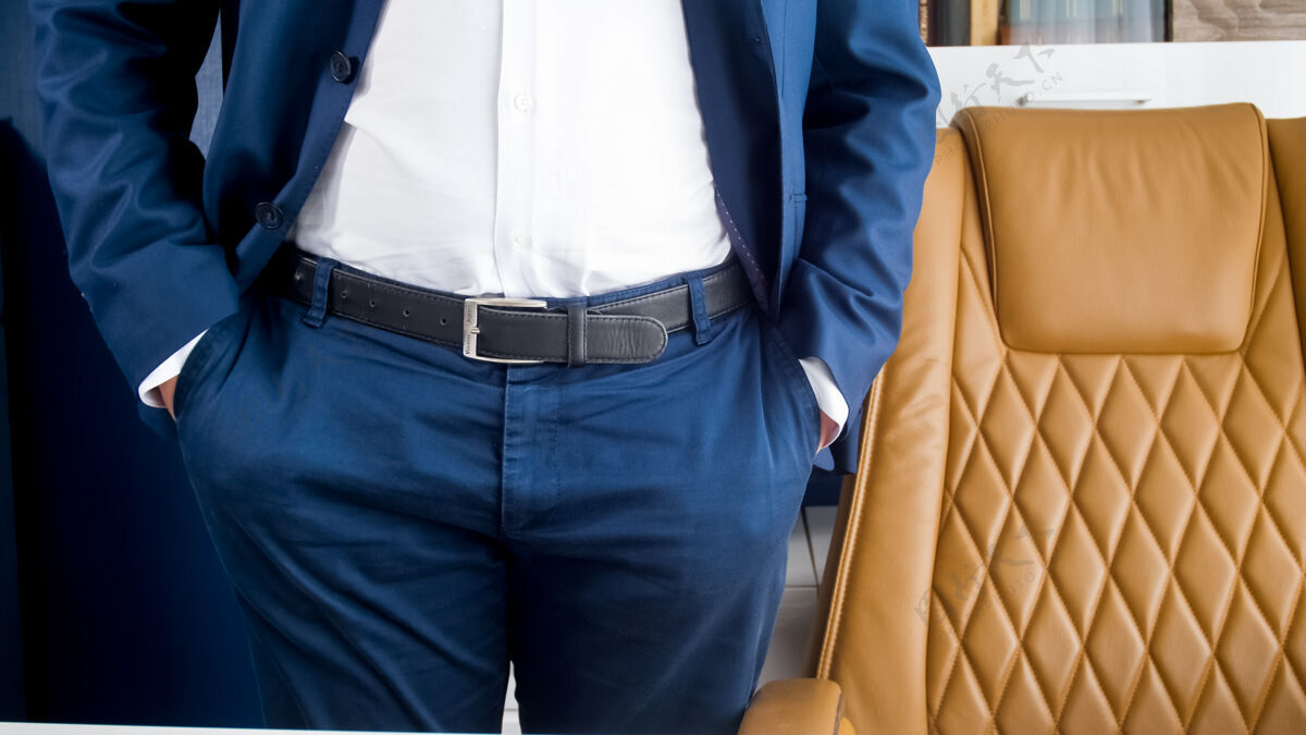 优雅穿着蓝色西装的时髦年轻商人站在现代化的办公室里 坐着棕色皮革扶手椅西装手表裤子