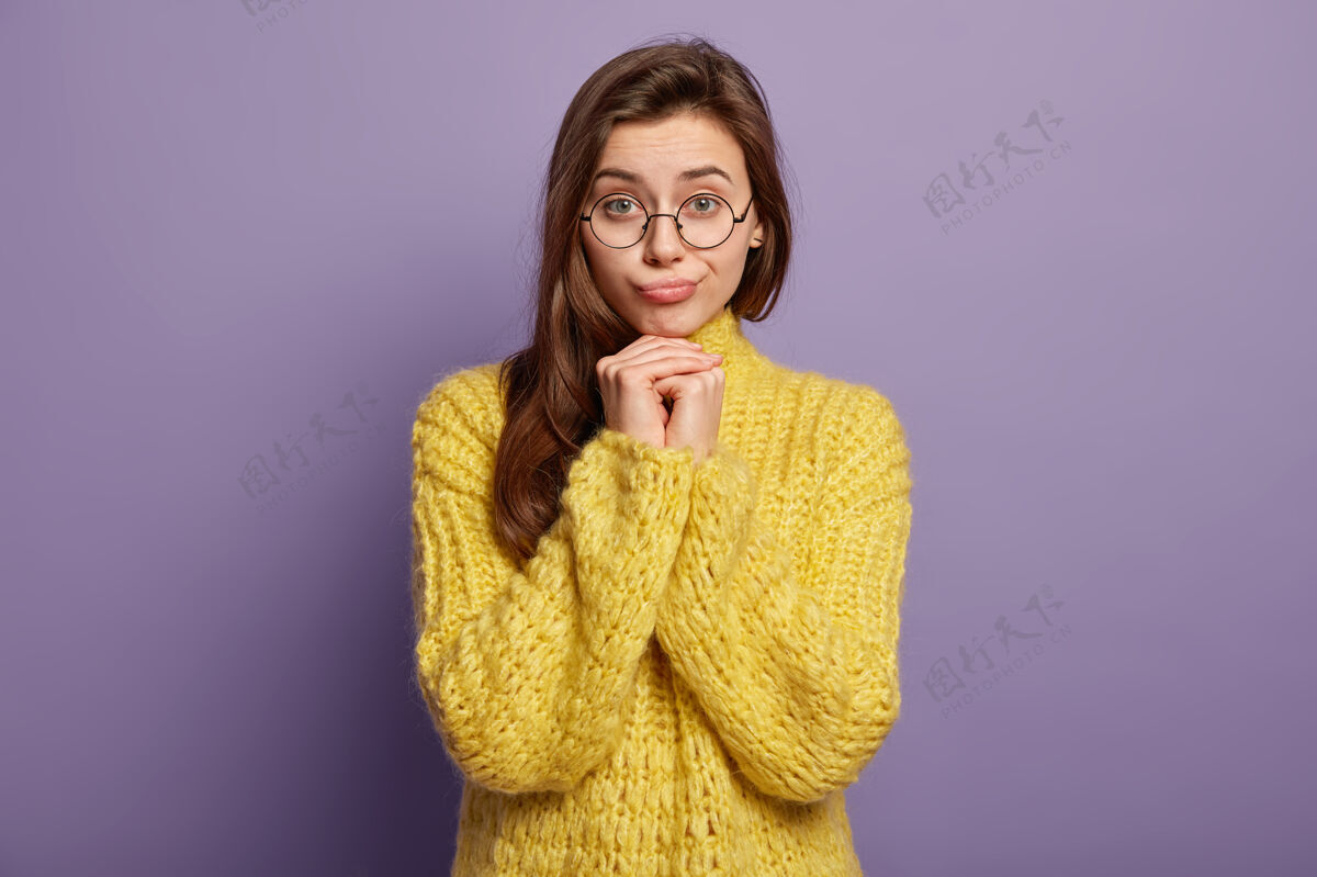 眼镜美丽的黑发女性的上身镜头感到孤独和悲伤 双手放在下巴下 被坏消息困扰 穿着黄色的套头衫 站在紫色的墙上人类的表情和感觉女人悲伤凝视