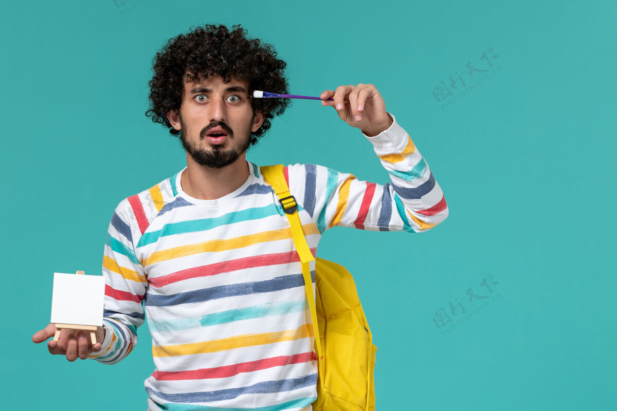 正面身穿条纹衬衫 背着黄色背包 拿着画架和流苏的男学生在蓝色墙上的正视图画架拿人