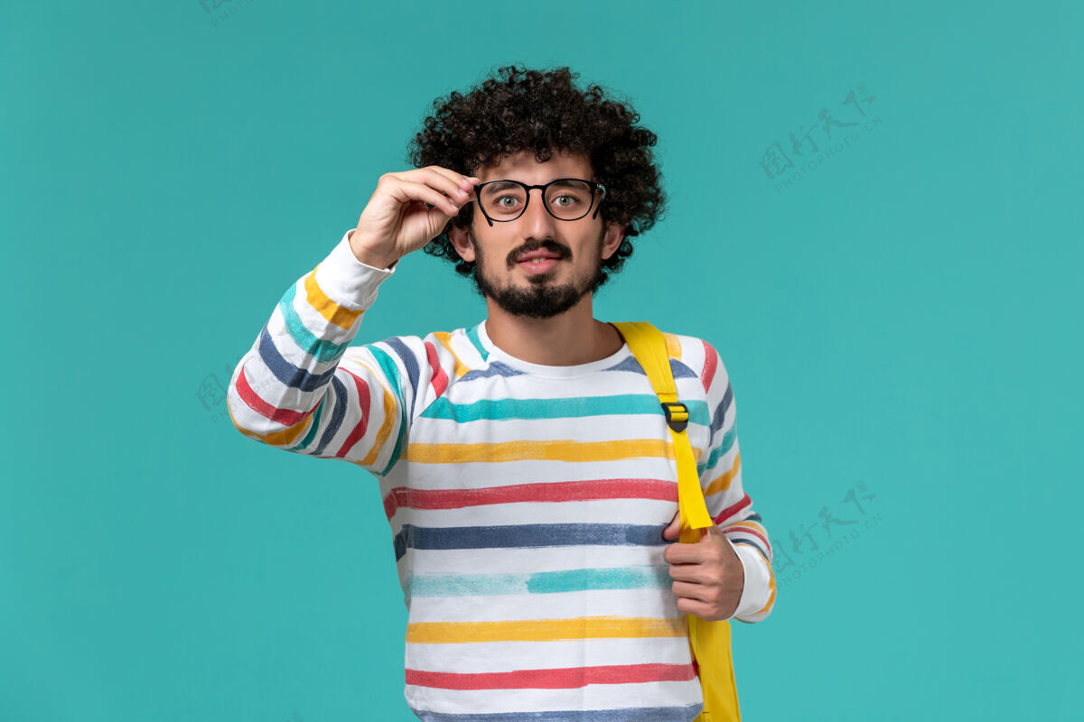 男性身穿条纹衬衫 背着黄色背包 手持光学太阳镜的男学生在蓝色墙上的正视图教训背包太阳镜