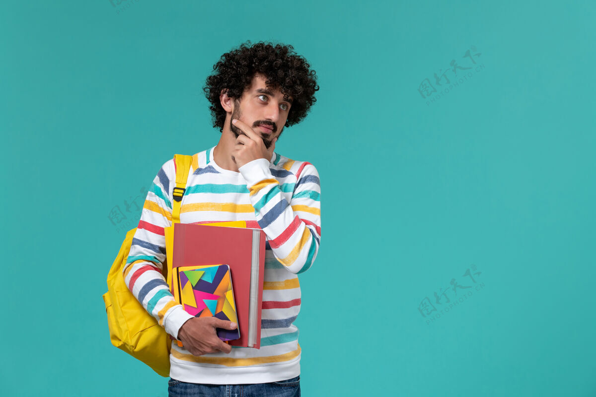 大学身穿彩色条纹衬衫 背着黄色背包 拿着文件和文案 在蓝色墙上思考的男学生的正视图背包大学条纹