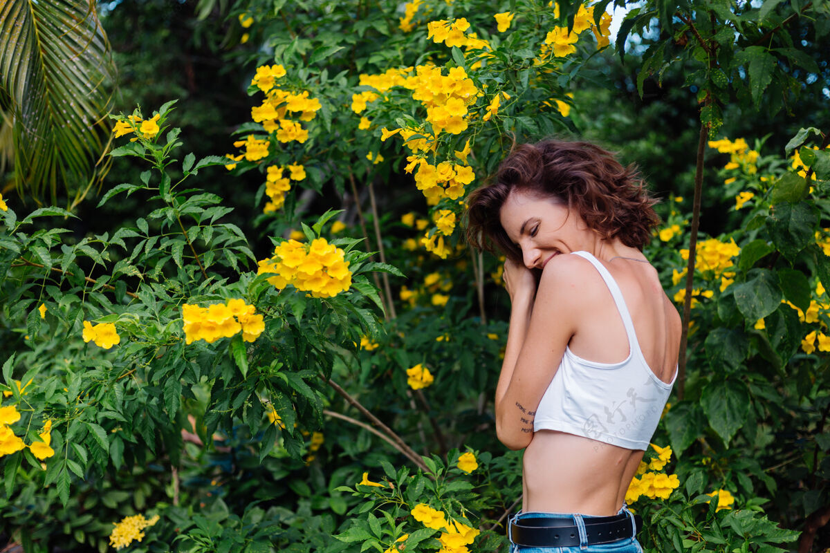 热带美丽的年轻苗条时尚合身的白人快乐女人 穿着白色短上衣和牛仔裤 公园里被黄色的泰国花包围花园女士公园