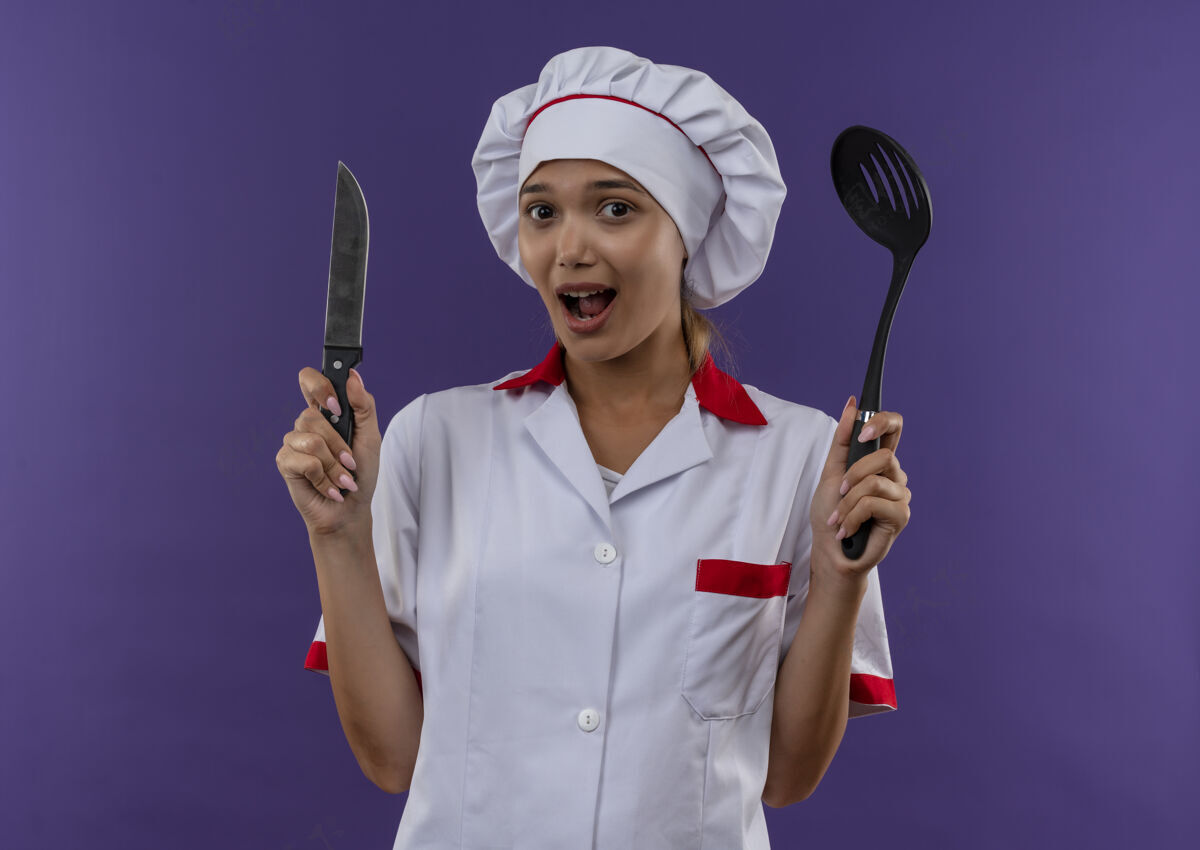 制服惊讶的年轻厨师女穿着厨师制服拿着勺子和刀在孤立的背景与复制空间拿着厨师女