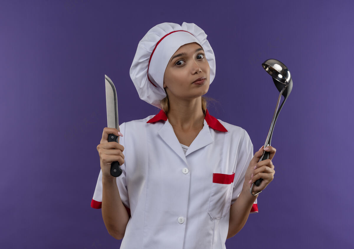 戴着年轻的女厨师穿着厨师制服拿着勺子和刀在隔离墙上厨师年轻刀