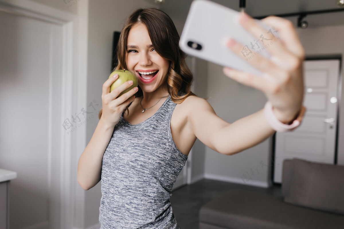 积极时尚女孩一边吃苹果一边拍照迷人女士用手机自拍的室内照片时尚自拍家