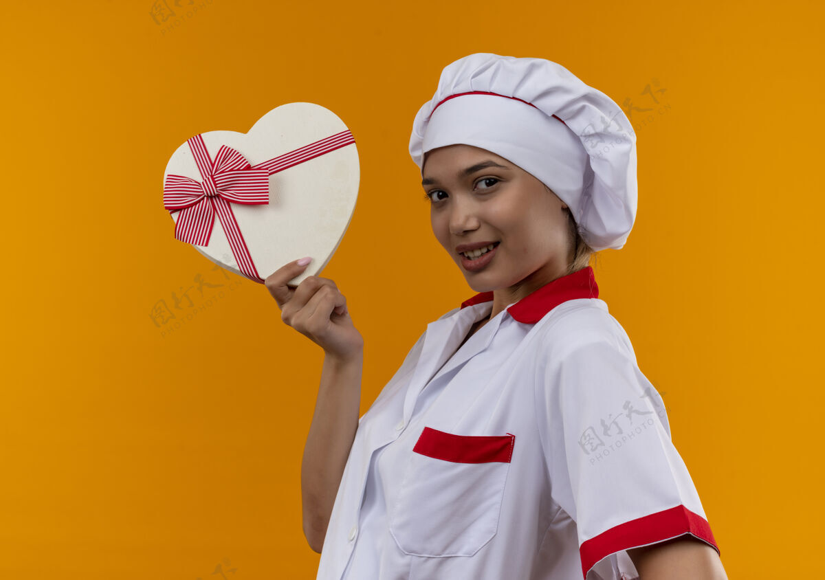 厨师高兴的年轻厨师女穿着厨师制服拿着心形盒子在孤立的橙色墙上高兴心女
