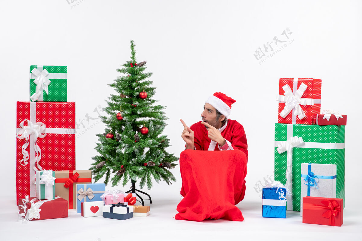 帽子一个悲伤的年轻人打扮成圣诞老人 拿着礼物和装饰过的圣诞树 在白色背景上指着右边的东西圣诞年轻人男人