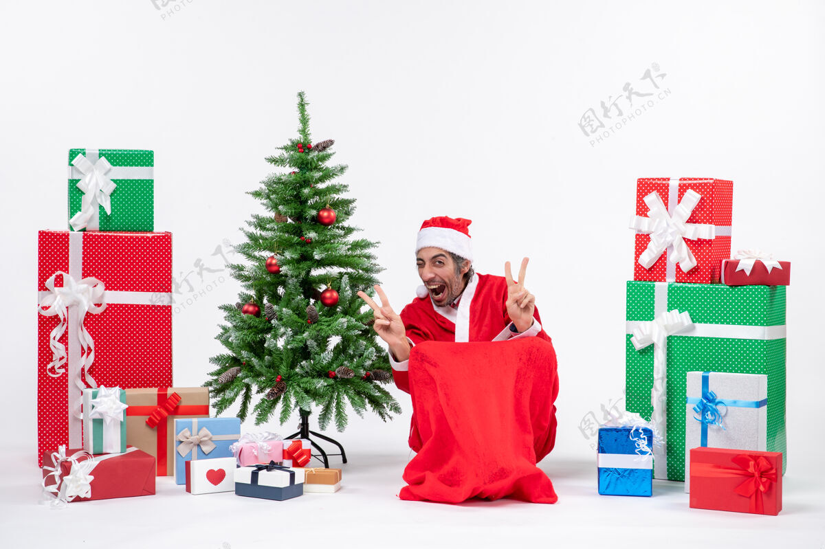 圣诞老人情绪激动的年轻人打扮成圣诞老人与礼物和装饰圣诞树作出胜利的姿态在白色背景年轻人打扮礼物