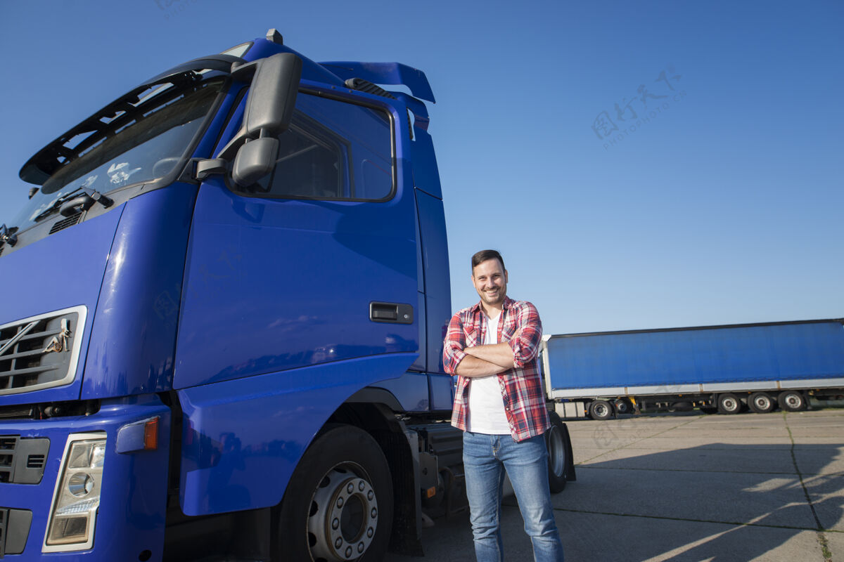 男卡车司机穿着便服站在他的卡车与双臂交叉在卡车站货物快速微笑